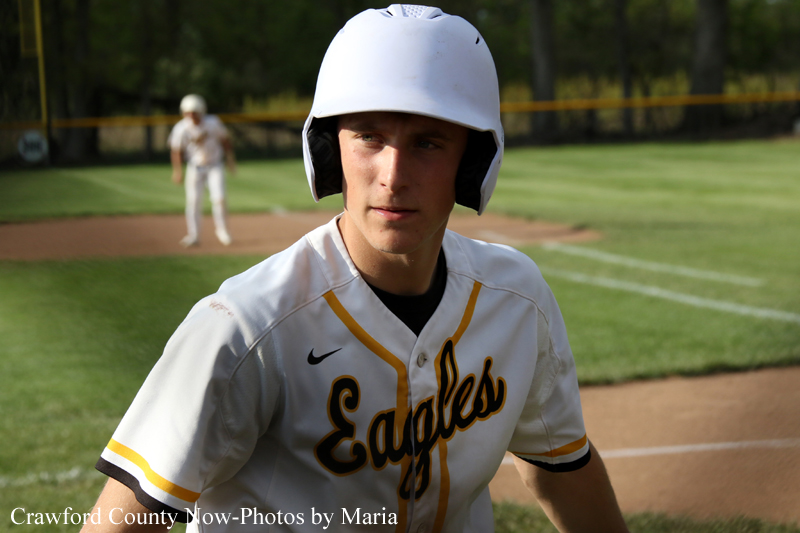 Baseball player in a white helmet and Eagles jersey stands on a sunlit field near home plate.