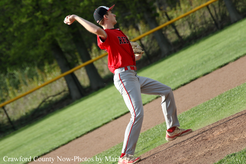 Baseball pitcher in a red uniform winds up to throw on the mound during a game on a sunny day.
