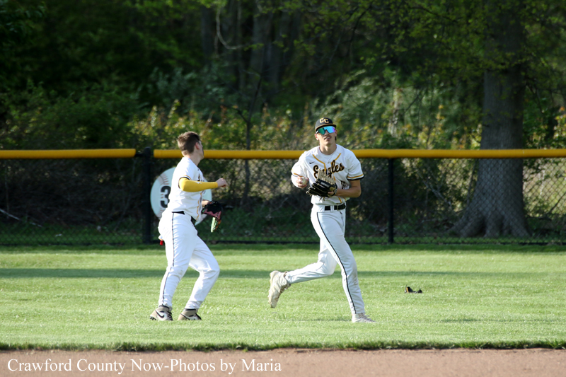 Two baseball players in white Eagles uniforms on a sunny outfield; one runs with a glove as the other prepares to field the ball.