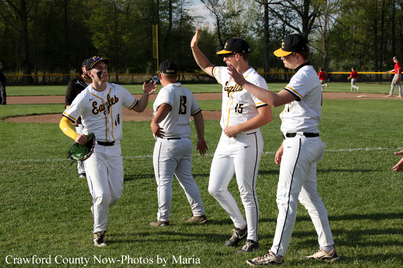 Four baseball players in white and yellow uniforms celebrate on a grassy field, giving high-fives after a game with a fence and outfield in the background.