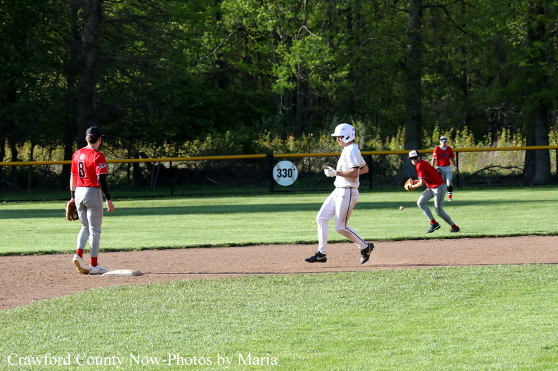 Baseball game on a sunny field with a runner in white approaching first base as a fielder in red watches nearby.