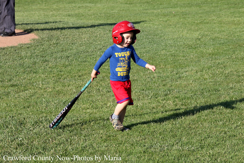 Young child wearing a red helmet, blue long-sleeve shirt and red shorts runs with a baseball bat on a grassy field.