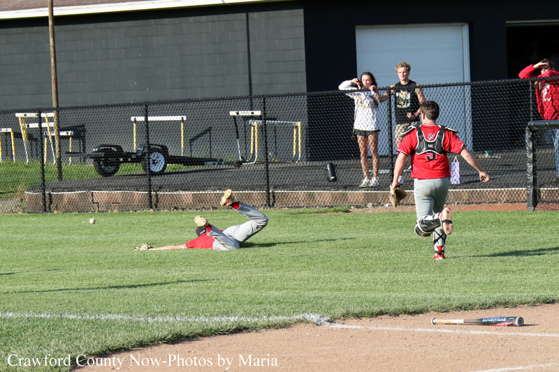 Baseball game: fielder slides on the grass to reach a ball while the catcher in red gear runs toward the play; spectators behind a chain-link fence.