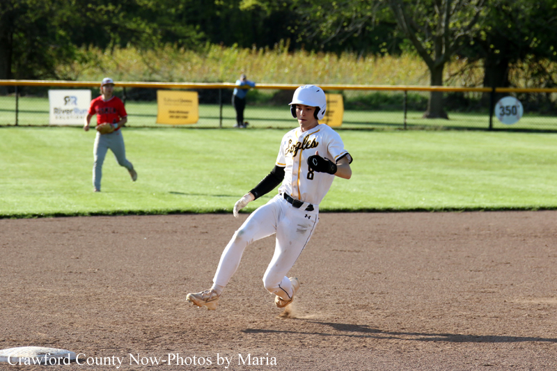 Baseball player in white uniform and helmet runs on the infield dirt during a game, with teammates and outfield signage in the background.