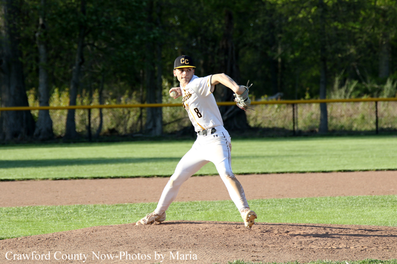 Baseball pitcher in white uniform releasing a pitch from the mound on a sunny field, trees in the background, cap with 'CC' logo. sports photo credit: Crawford County Now—Maria.