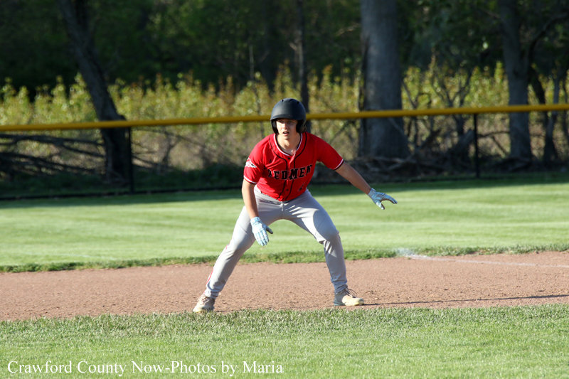 Baseball player in red jersey crouches on the infield, ready to field a ground ball on a sunny day.