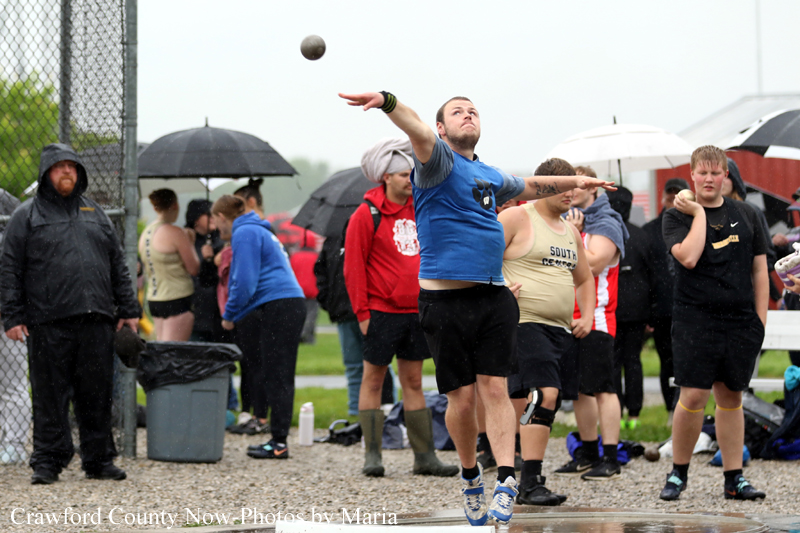 Athlete in a blue shirt launches a shot put during a rainy track event, crowd with umbrellas watches in the background.