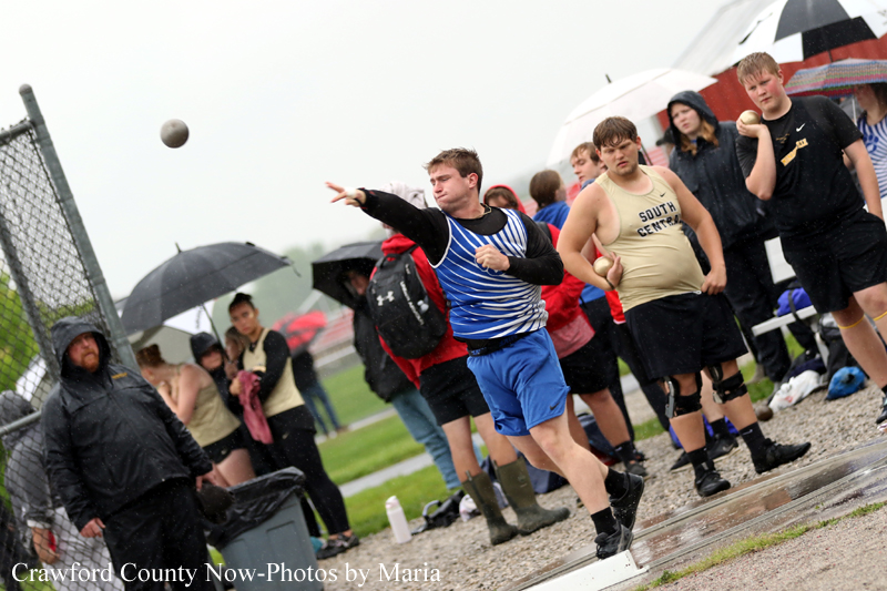 Male shot-put athlete in blue striped top and blue shorts throws a ball on a wet track as spectators with umbrellas watch in the rain.