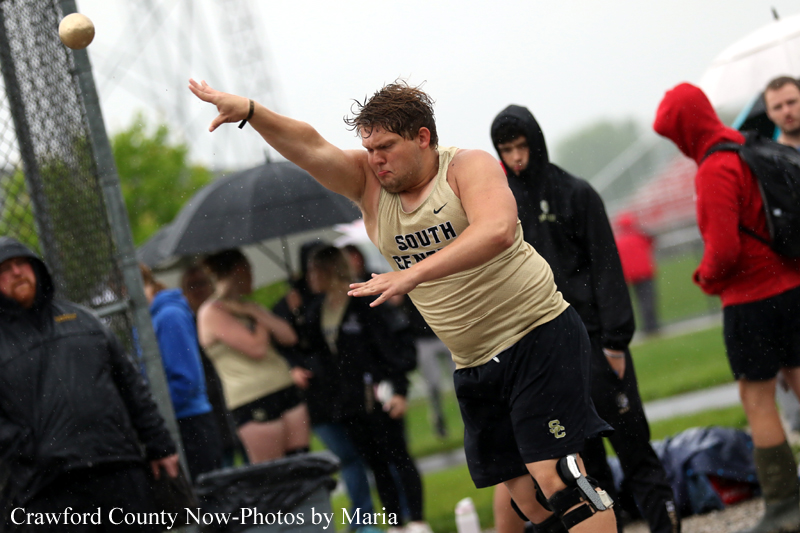 Male track and field athlete in a beige singlet mid-throw of a shot put on a rainy day, with spectators under umbrellas in the background.