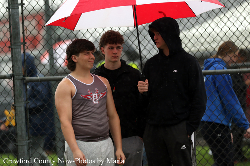 Three teens stand by a chain-link fence in the rain; one wears a gray sleeveless shirt, another in a black hoodie holds a red-and-white umbrella for cover.
