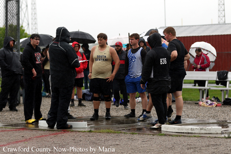 Group of young male runners in starting-position gear listen to a coach on a rainy day at the track, with wet blocks and spectators under umbrellas in the background.