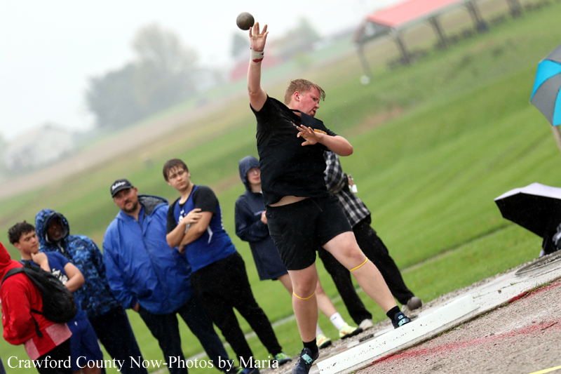 Male athlete in black throws a shot put during a track and field event while spectators watch on a grassy field in the background.