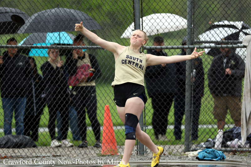 Female athlete from South Central in a beige jersey and black shorts mid-throw/landing pose during a long jump in a rainy meet, with spectators behind a chain-link fence and umbrellas.