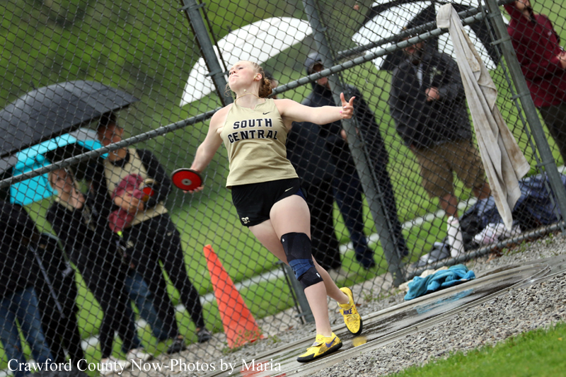 Female athlete in a beige 'South Central' shirt throws a discus during a track meet, behind a chain-link fence with spectators watching under umbrellas.