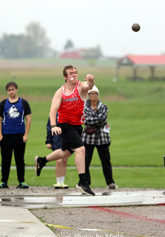 Athlete in red singlet throws a shot put on a track field as spectators watch in the background, outdoors on a cloudy day.