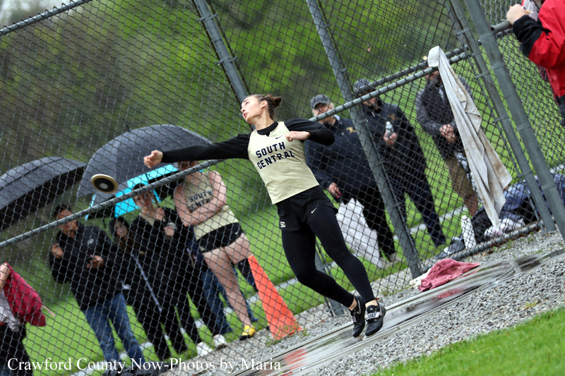 Female discus thrower in a cream and black uniform releases a throw near a chain-link fence while spectators watch behind her on a grassy field.