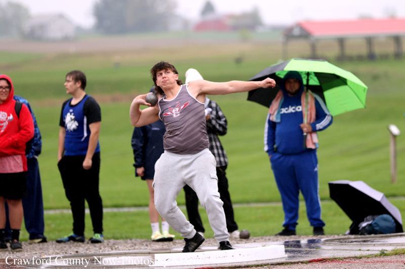 Shot-put athlete mid-throw in a gray tank top on a wet track, with onlookers in the background, some under umbrellas in the rain.