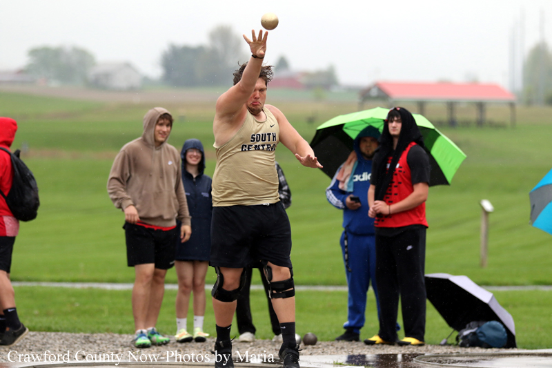 Male shot-put athlete mid-throw on a wet track, spectators under umbrellas watching.