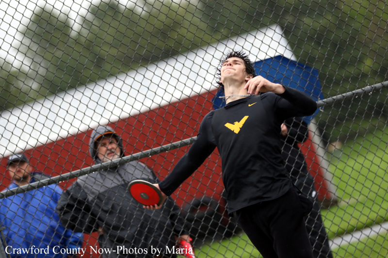 Male discus thrower in a black athletic shirt releases a red discus at an outdoor meet, chain-link fence in the foreground.