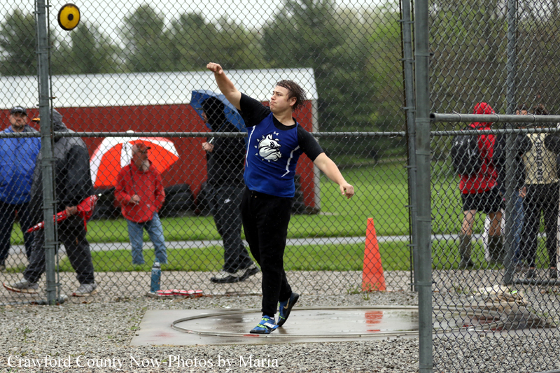 Male athlete in a blue jersey throws a yellow discus inside a fenced throwing circle at a track and field meet with spectators nearby.