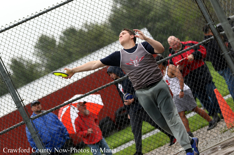 Discus thrower mid-throw reaching forward with a discus behind a chain-link fence, with spectators watching nearby far side of the field.