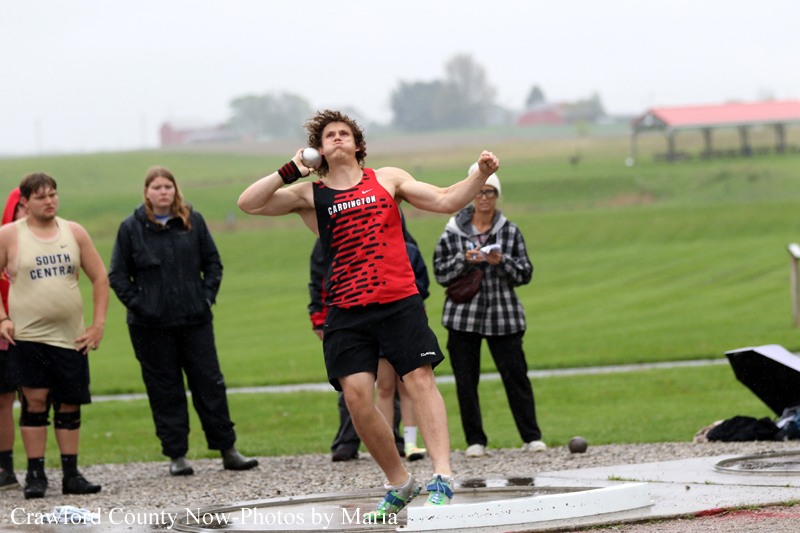 Male athlete in red and black uniform mid-throw of a shot put, spectators watching on a grassy field behind him.
