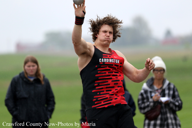 Male athlete in a black and red Cardington singlet is mid-throw with a shot put on a grassy field, spectators watching in the background.