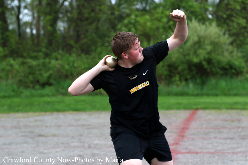 Man in a black shirt mid-shot put throw on an outdoor field, ball near his neck, with greenery in the background