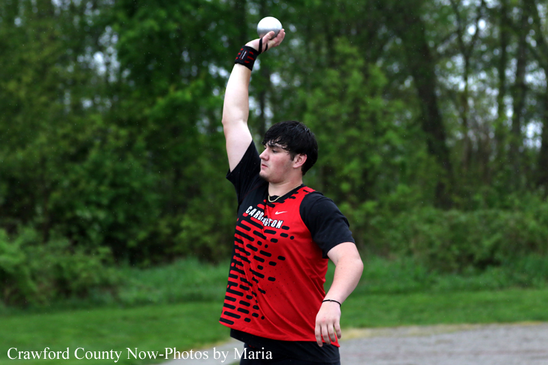 Male athlete in a red and black uniform throwing a shot put outdoors on a grassy field with trees in the background.