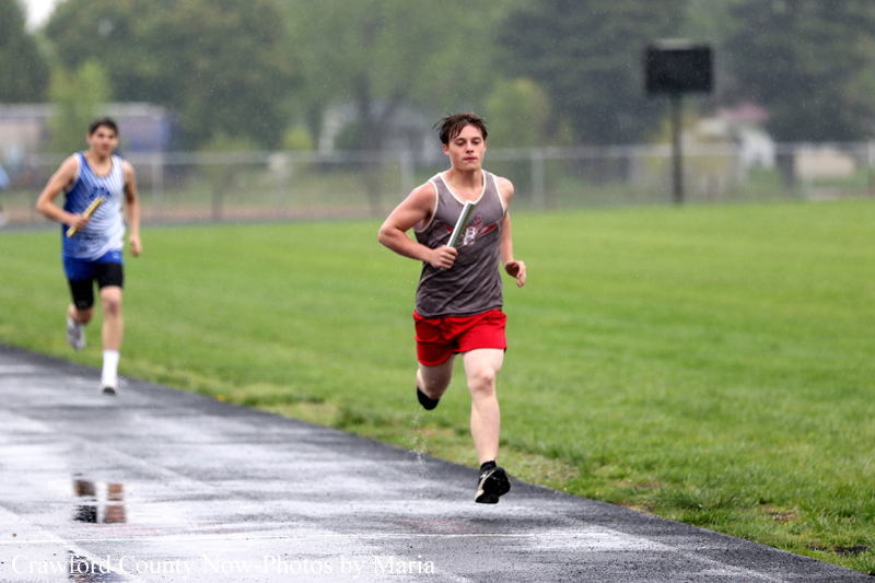 Male sprinter in a gray sleeveless shirt and red shorts holds a baton while sprinting on a wet track; a second runner in blue stripes follows behind in the rain.
