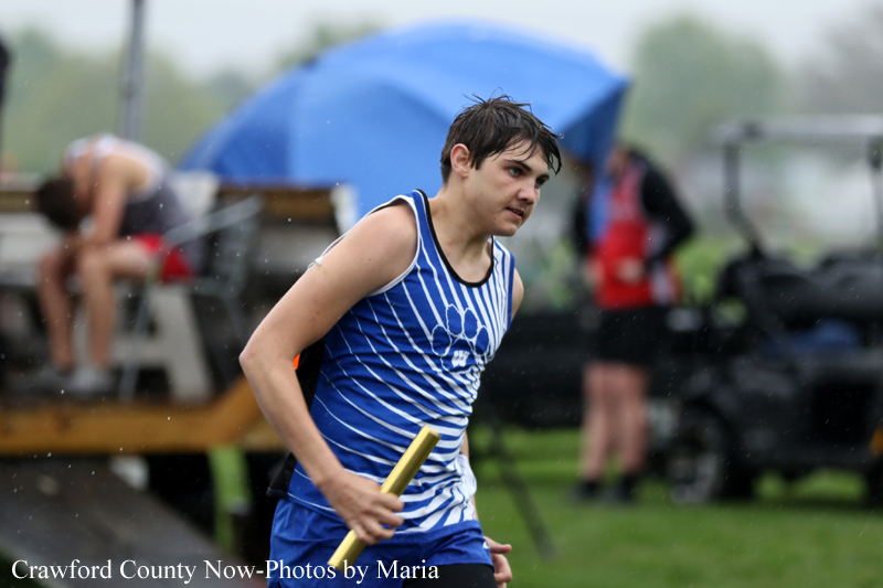Young male relay runner in a blue striped uniform grips a baton while sprinting outdoors in rainy weather, blurred background athletes nearby.