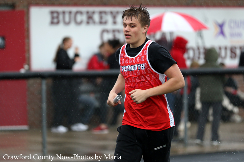 Male athlete in a red Plymouth uniform runs with a baton during a relay on a wet track, with spectators in the background.