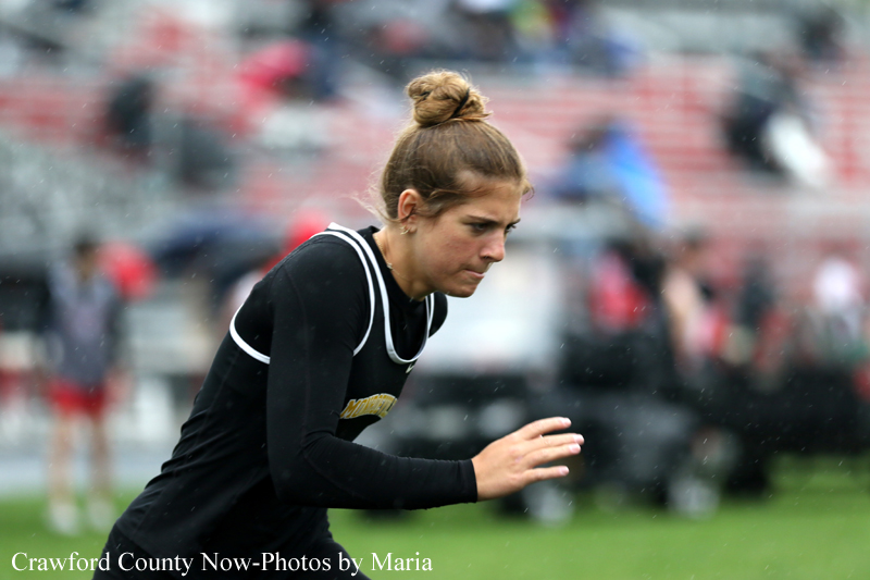 Female sprinter in a black sleeved jersey running on a track with blurred stadium seats behind her, focused on the race.