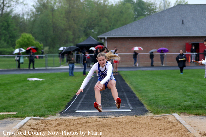 Female athlete in mid-air performing a long jump over the sand pit during a rainy track meet, spectators with umbrellas in the background.