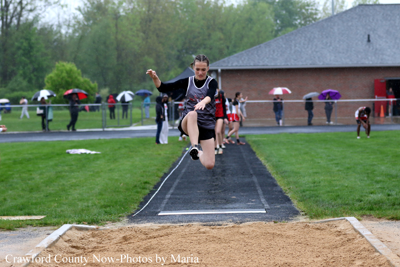 Female athlete mid-jump in a long jump at a track meet, just above the runway with the sand pit ahead and spectators with umbrellas in the background.