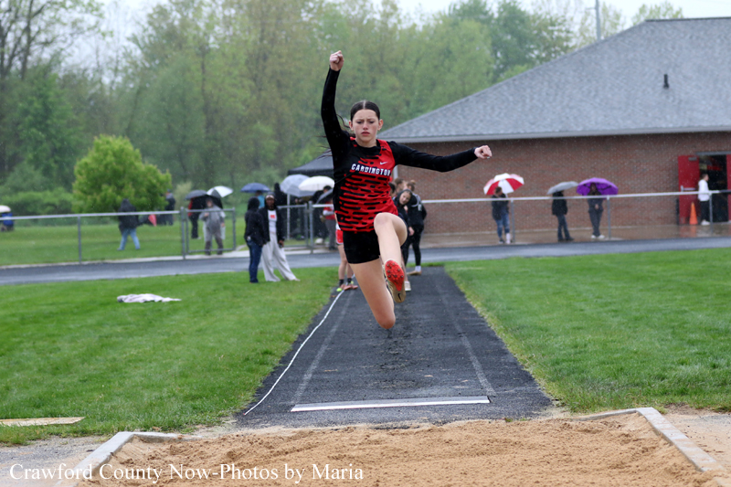 Female athlete in red and black uniform mid-air during a long jump toward the sand pit at a rainy track meet.