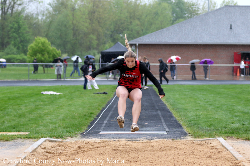 Female athlete in red uniform mid-jump at the long jump pit, sand ahead, spectators with umbrellas in the background.