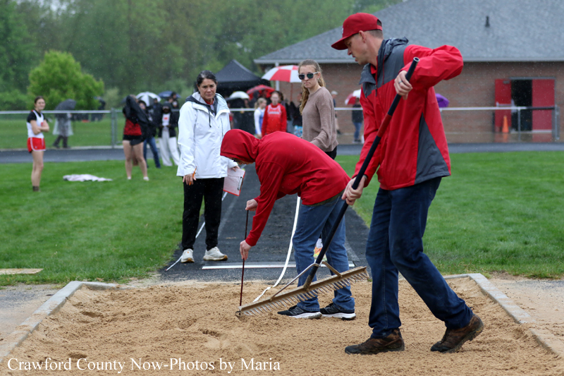 People at a track field preparing a sand pit for a long jump, with a coach raking the sand as a student helps level it in the rain.