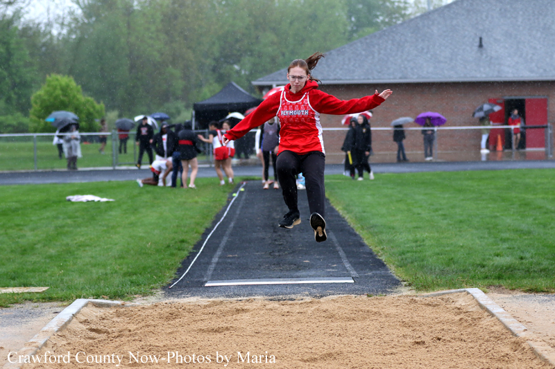 Female athlete in a red shirt mid-air during a long jump at a track meet, sand pit in foreground and spectators behind on a rainy day.