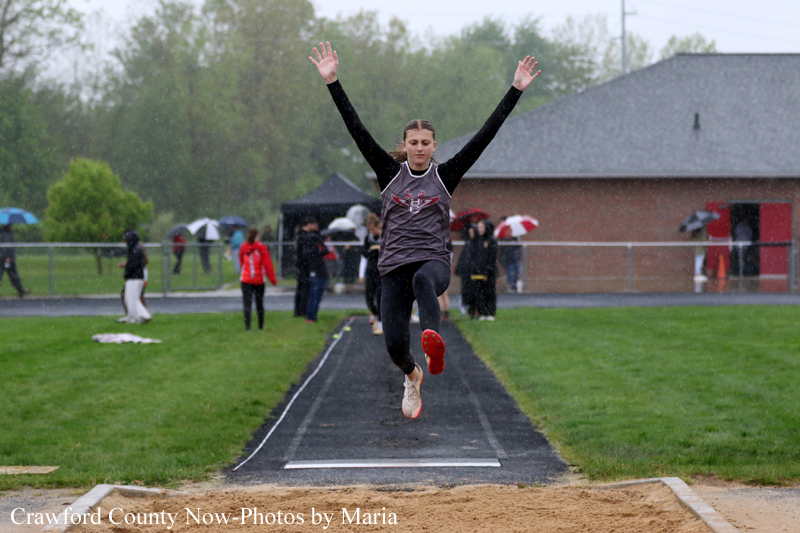 Female athlete mid-air performing a long jump in the rain, landing area ahead, spectators with umbrellas in the background.