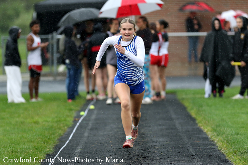 Young female athlete sprinting on a wet track during a race, spectators with umbrellas watching in the background.