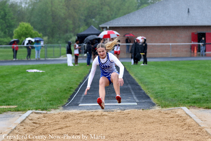 Female athlete in blue and white uniform mid-air performing a long jump, over the sand pit as spectators with umbrellas stand by in the rain behind the track.