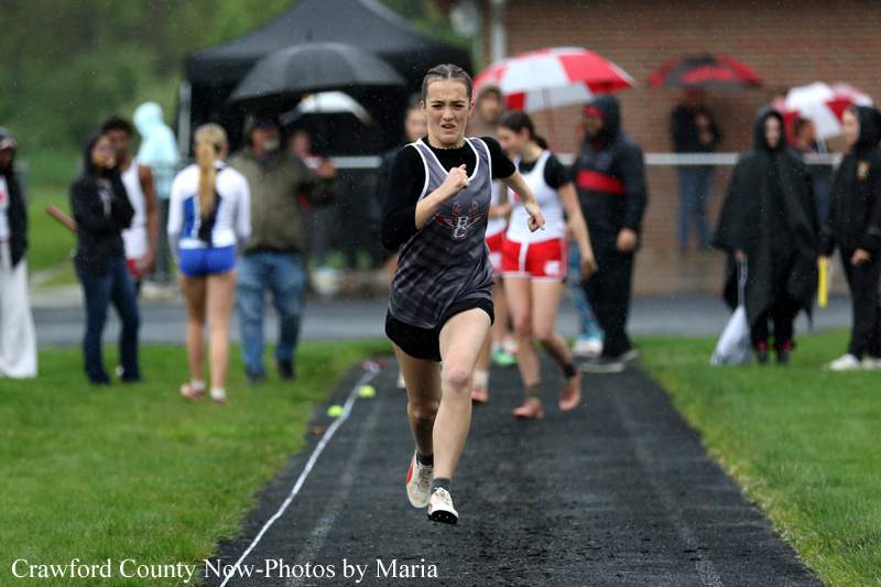 Female sprinter in gray-black uniform runs on a wet track; spectators with umbrellas in the background.