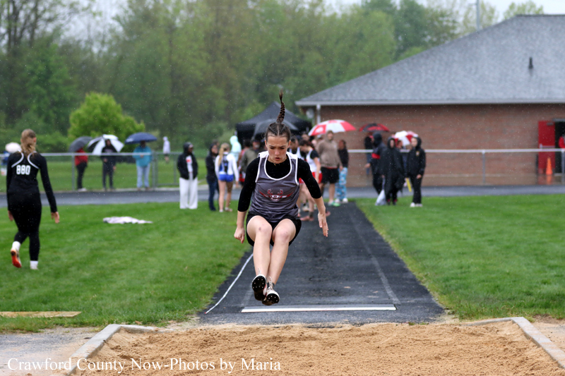 Female long jumper in midair during a track and field event, about to land in the sand pit as spectators watch.