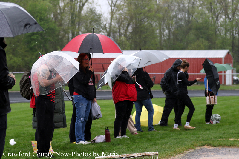 People stand in a line on a grassy field holding umbrellas in the rain, facing left to right.