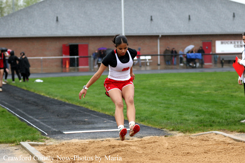 Female track athlete in red shorts and white top lands in the sand pit during a long jump, with a track and onlookers in the background