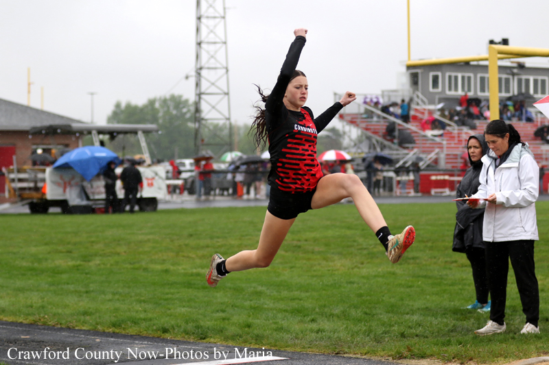 Female athlete mid-air performing a long jump at a track meet on a rainy day, with officials nearby watching. (Informative)