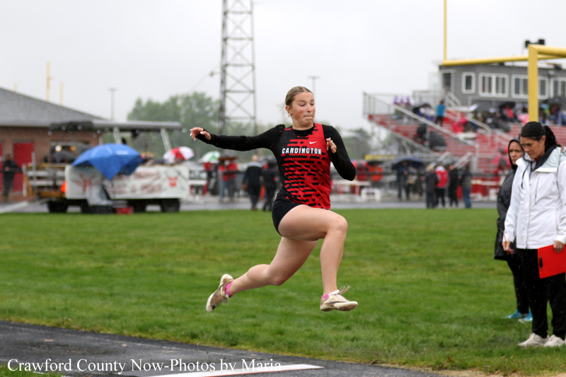 Female athlete in a red and black Cardington uniform mid-air during a jump on a rainy track, with spectators and umbrellas in the background.