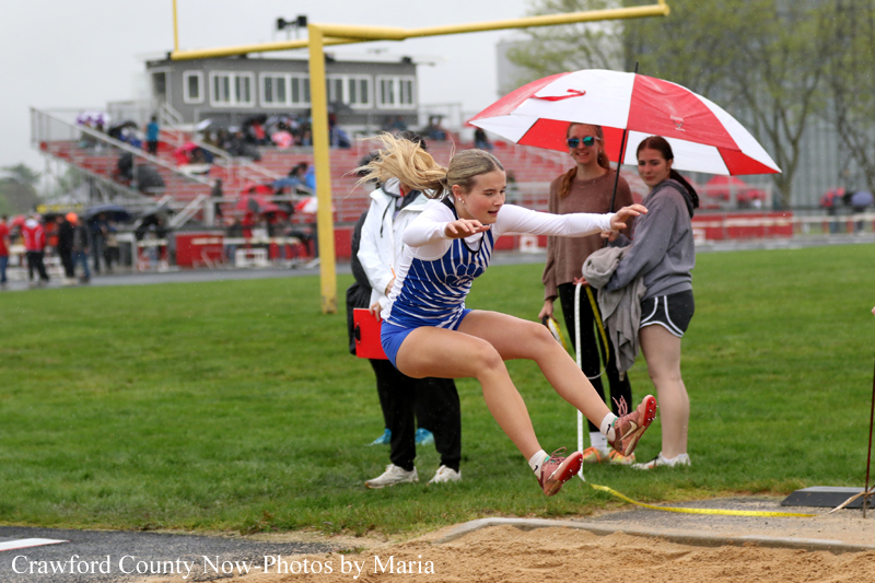 Female athlete in blue and white uniform mid-air during a long jump, landing in the sand pit as spectators watch nearby under an umbrella.