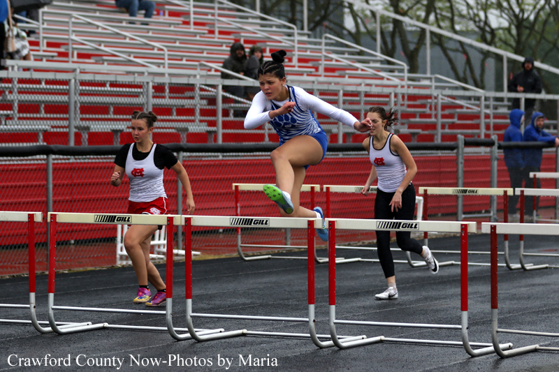 Three female athletes sprint over hurdles on an outdoor track, the middle runner mid-air clearing a barrier as two others approach. Red bleachers and a chain-link fence backdrop a rainy day stadium.